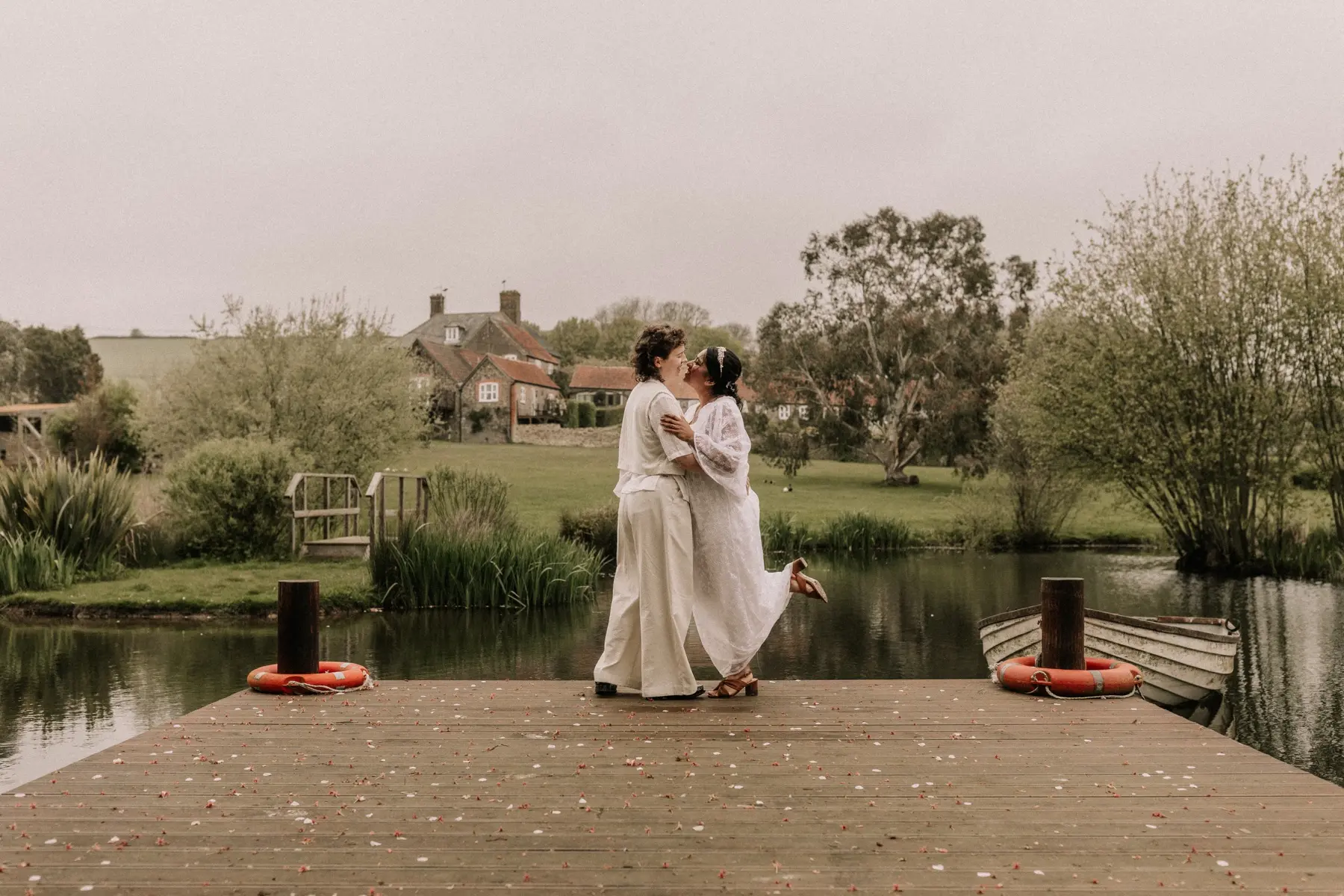 Two brides embracing beneath a wooden oak arbour reflected in still water at Rudge Farm
