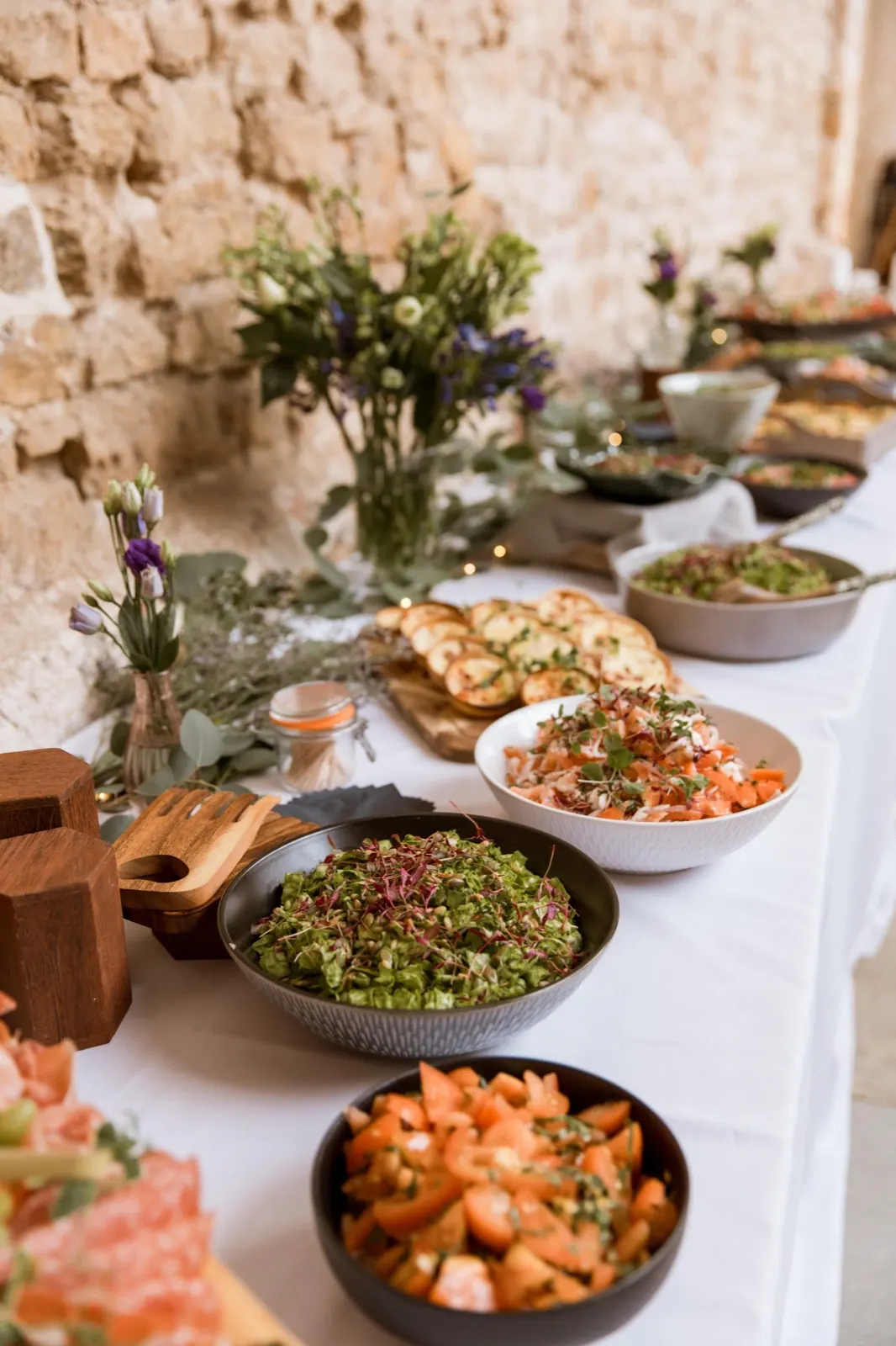 A long table of sharing food by Ako Dining against a stone wall, bowls of salad and flowers down the middle