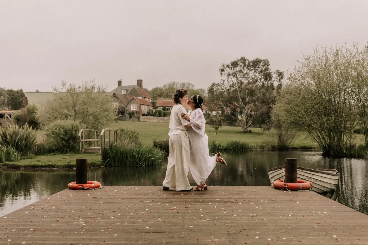 Two brides kissing on a wooden jetty over a still lake at Rudge Farm, the farmhouse and Dorset countryside behind them