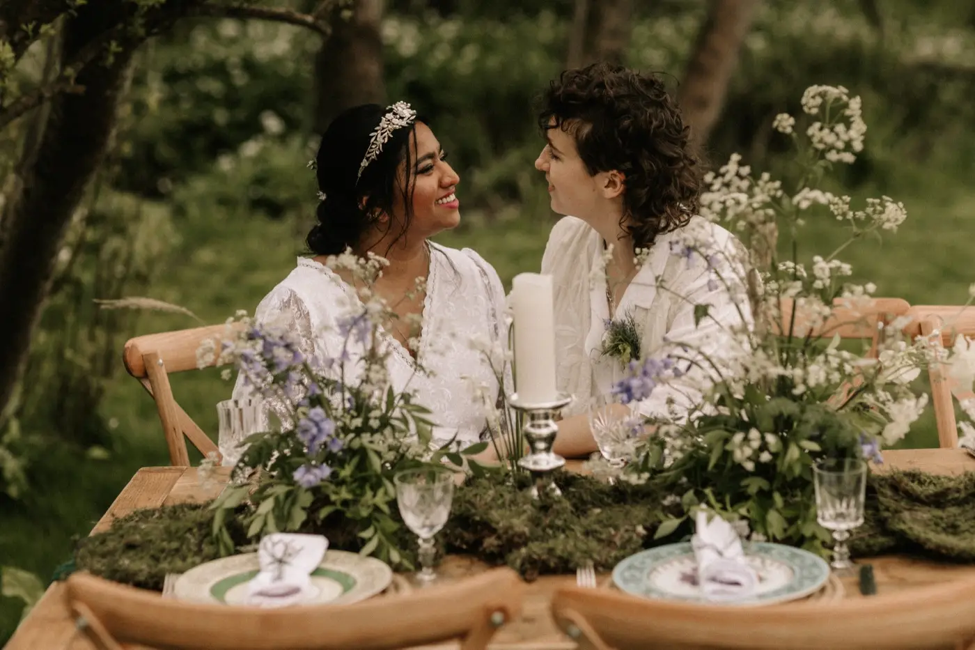 Nadia and Poppy at the woodland table, smiling at each other across the flowers and candles
